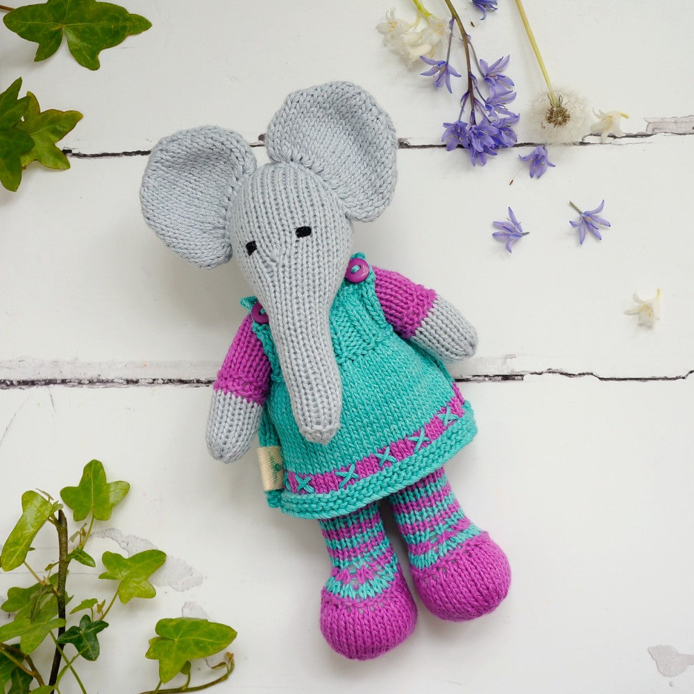 Hand-knitted grey elephant wearing a green-blue dress, striped leggings and purple-pink shoes on a white background with fresh flowers and foliage.
