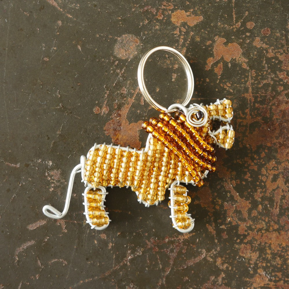 A handmade lion-shaped keychain crafted with gold and white glass beads and galvanised wire, resting on a textured dark brown surface.