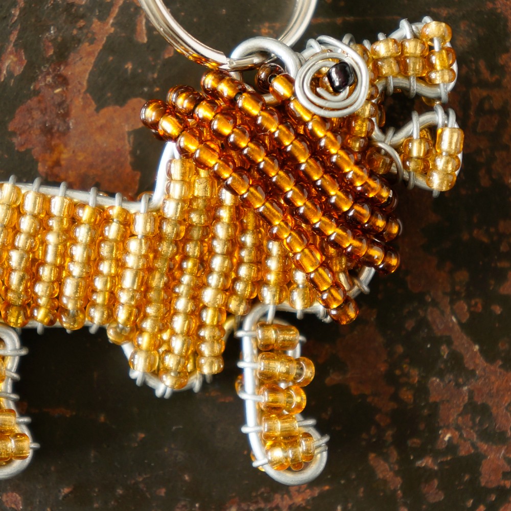 A handmade beaded lion keychain made from amber and gold glass beads and galvanised wire, shown close-up on a dark textured background.