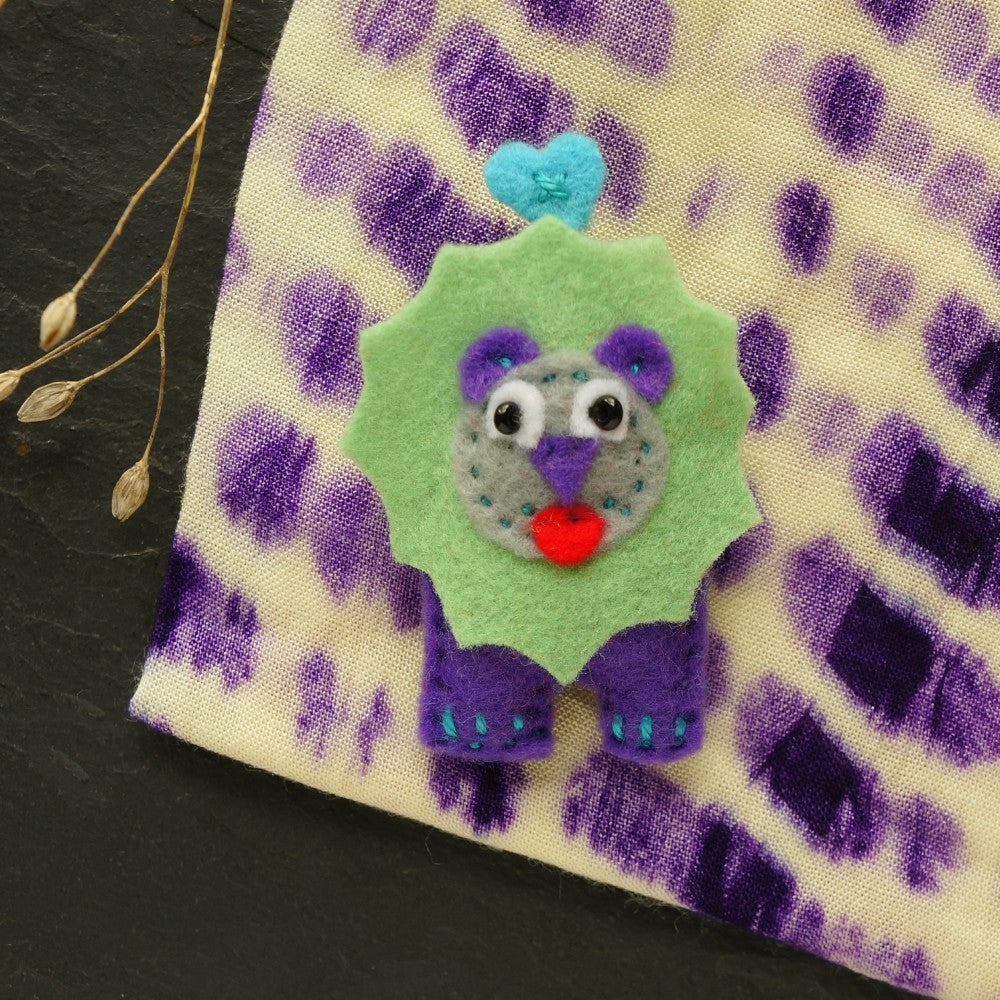 Felt lion brooch with green mane, purple body, cheeky red tongue on spotted purple fabric background