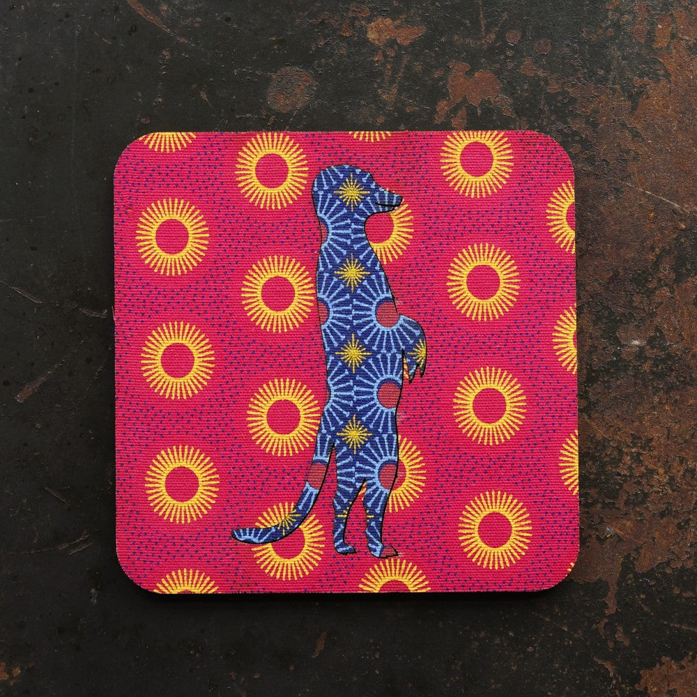 Coaster with a blue meerkat illustration on a pink background with yellow sun patterns, placed on a dark surface.