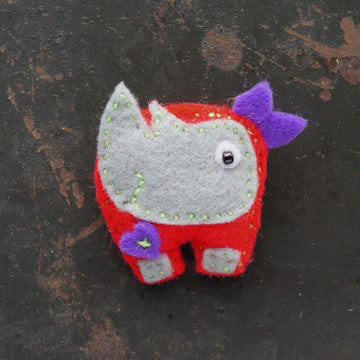Small felt rhino brooch with red body, grey face, and purple ears on a dark background