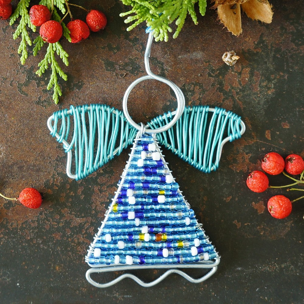 A handmade wire angel Christmas ornament with a blue beaded body, decorated with small colorful beads, hanging against a rustic dark brown background with red berries and green pine branches.
