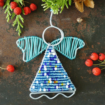 A handmade wire angel Christmas ornament with a blue beaded body, decorated with small colorful beads, hanging against a rustic dark brown background with red berries and green pine branches.