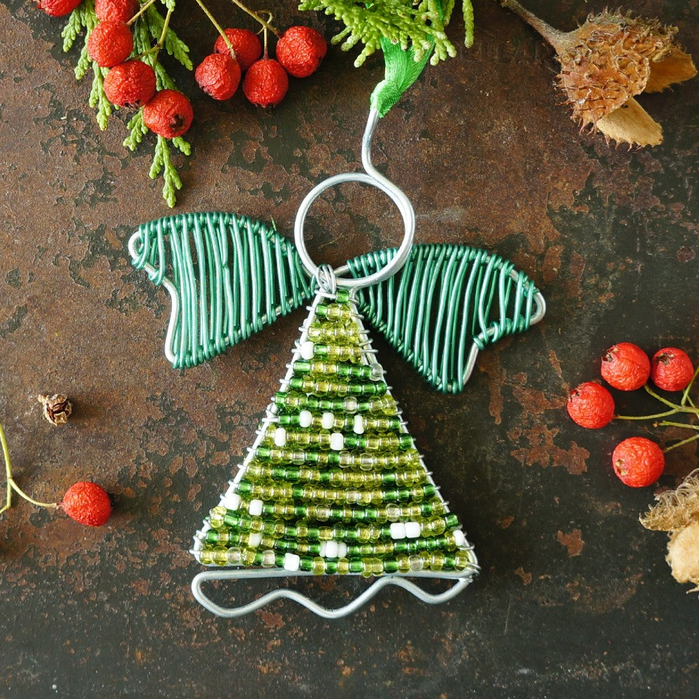 A handmade wire angel Christmas ornament with a green sequined body, green wire wings styled on a rustic dark metal surface with red berries and dried botanicals.
