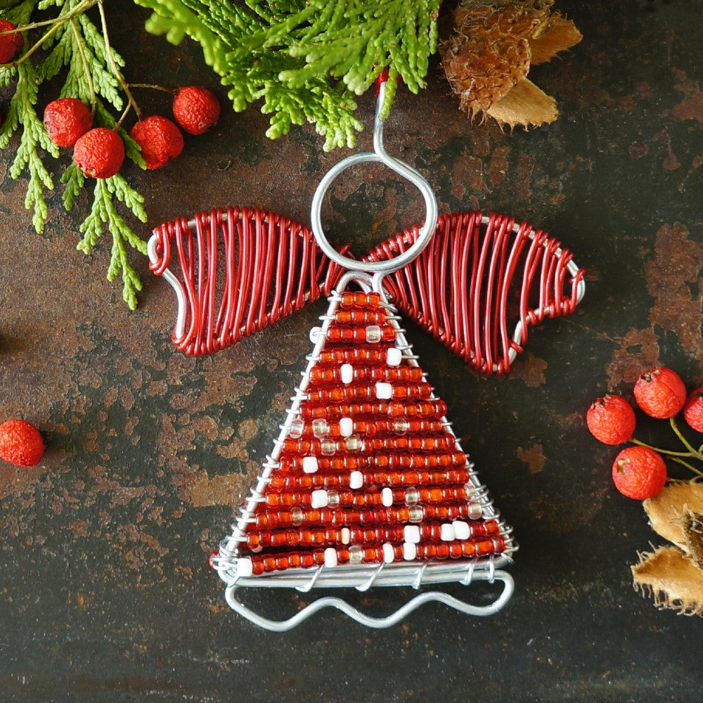 A handmade Christmas angel ornament made from red wire wings and a red beaded tree body with white bead accents, displayed on a rustic dark background with evergreen branches and red berries.
