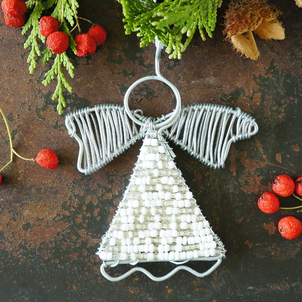 A handmade wire angel Christmas ornament with a silver wire-wrapped wings and body decorated with white beads, displayed on a rustic dark brown surface with holiday greenery and red berries.
