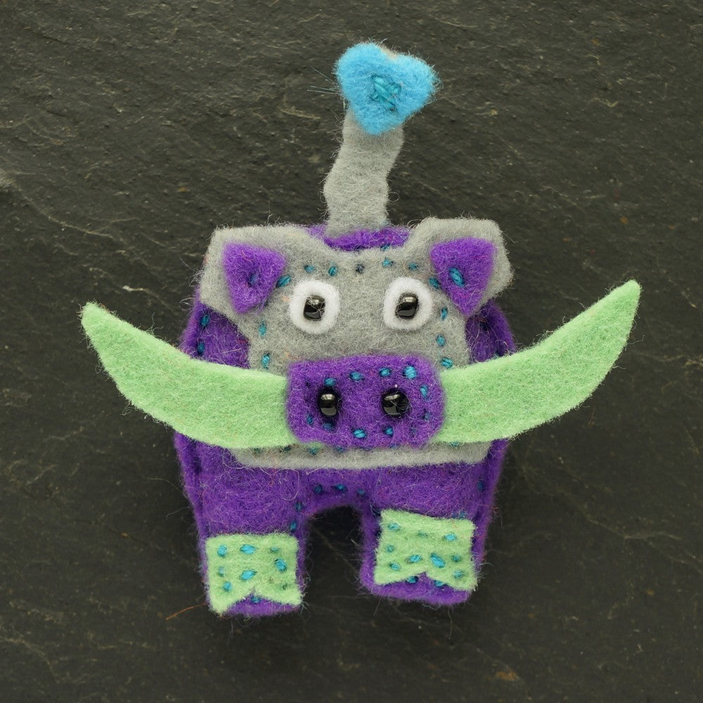 Felt warthog  brooch with purple body, green tusks, and grey face on a dark background